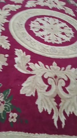 This video captures a close-up, angled view of a large, ornate rug with a deep red background. The rug features a complex pattern of cream-colored floral and scrollwork motifs, along with clusters of pink and green roses and smaller white flowers scattered across its surface. A cream-colored border with a zigzag pattern frames the edge of the rug. The camera pans slowly across the rug, highlighting the intricate details of its design. The lighting is somewhat dim, creating shadows that emphasize the texture and depth of the rug.