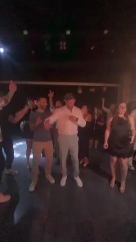The video shows a group of people dancing in a dimly lit club with purple and pink lights. Most people are wearing casual clothing. A man in a white shirt and light-colored pants is in the center, dancing. A woman in a black dress is to his right. A man in a striped shirt is on the left, also dancing. There are several other people dancing in the background. The atmosphere appears to be celebratory.