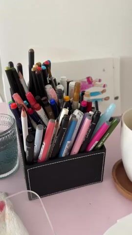 A close-up shot focuses on a black rectangular container filled with an assortment of writing utensils, including pencils, markers, and pens of various colors and sizes. The container sits on a light pink desk next to a textured glass of water and a wooden coaster topped with a white mug. Behind the container, a white board is visible, displaying a simple sketch of figures. A hand wearing a lace glove reaches into the container, shuffling through the items before extracting a black pencil.