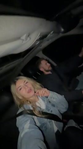 A young woman in a denim jacket and a man driving are in the front seats of a car at night, moving to music. The woman dances in her seat, singing along to the song, and the man occasionally sings as well.