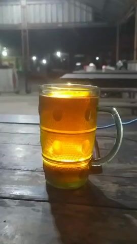 A close-up shot focuses on a glass beer mug filled with amber liquid, likely beer. The mug has a handle and a textured design around its body, with circular indentations that are illuminated from within by a yellow light, creating a glowing effect. The mug sits on a wooden surface, possibly a table, which has visible grain and texture. In the background, an outdoor area is dimly lit by various lights. Several light fixtures are visible, some appearing to be overhead. The environment suggests a public space like a restaurant patio or an open-air market at night. There are no people clearly visible in the frame, and no direct action is taking place other than the static presence of the illuminated beer mug.