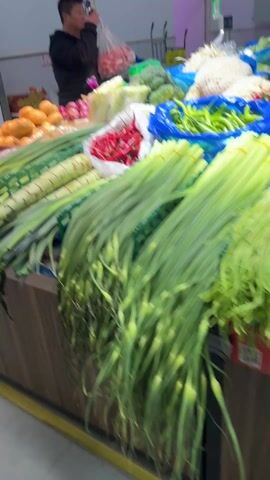 The video shows a vegetable and insect market stall, with an individual standing behind the stall looking at their phone and holding a plastic bag with red onions.  The stall displays a variety of produce, including green onions, potatoes, and red peppers, but the prominent feature is several large metal trays filled with what appear to be cooked or fried silkworm pupae. There are also some chestnuts in a separate tray. A phone number is visible on the side of the stall.