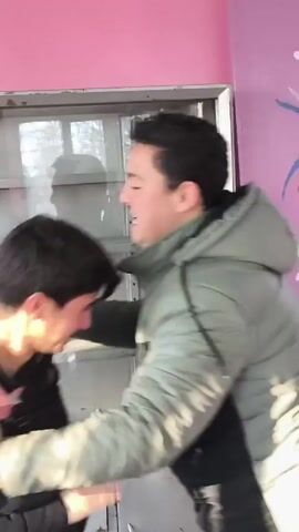Two young men playfully wrestle outside, one holding what appears to be a lollipop in his mouth. They stumble and jostle each other into a building with vividly colored walls. Inside, two other young men join the playful pushing and shoving around a low, cage-like structure with a mesh top.