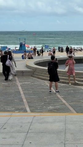 The video opens with a view of a beach scene. A man wearing a black t-shirt, black shorts, and a black backpack is standing on a paved area, holding a phone and taking a picture of a woman. The woman, wearing a pink floral dress and a straw hat, is standing on stone steps and posing for the photo. Behind them, there is a large bronze sculpture depicting a stylized figure. In the background, the ocean is visible with waves and a hazy sky. The beach is populated with many people, some sunbathing, others walking or standing. There are umbrellas and beach chairs on the sand. A blue lifeguard stand is also visible on the beach. As the video progresses, the camera pans slightly to the right, revealing more of the sculpture and the people around it. Two more individuals, a man and a woman, are seen interacting near the sculpture, with the woman adjusting the man's collar. Other people are visible on the steps and in the background, enjoying the beach. The overall environment is a public beach area with a prominent sculpture, suggesting a tourist destination. The footage is shot during the daytime under an overcast sky.
