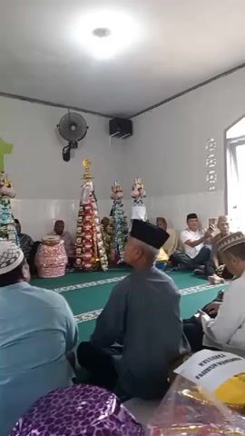 The video shows an indoor gathering, likely a mosque, with several people sitting on a green carpet. In the center of the room, there are several tall, cone-shaped displays decorated with various items, possibly food or gifts. A man is standing on a raised platform, holding a microphone and appearing to speak. Other individuals are seated on the floor, some facing the speaker and others observing the displays. Some people are wearing traditional Islamic attire, including skullcaps. The walls are white with some green architectural elements. There are also fans mounted on the walls. The overall atmosphere appears to be one of a religious or communal event.