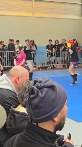 In a brightly lit indoor space, an amateur kickboxing match unfolds on a blue mat. Two competitors wearing protective headgear, one red and one yellow, engage in combat. The athlete in red lands a kick on the yellow helmeted competitor. They both wear black T-shirts, shin guards, and dark socks. The fighter in red also wears gray shorts, while the other wears patterned shorts.

The scene is observed by spectators standing behind a blue metal barricade that rings the ring.  Some wear black T-shirts with a circular logo on them. The audience includes a person in a pink shirt with the word “SILVERT TEAM” printed on it, as well as a man in a black beanie in the foreground, with his face partially visible. Others wear various clothing items like jackets, hoodies, and sweatpants.  

The venue has white walls and a high ceiling with fluorescent lighting. An exit sign is visible on the wall behind the crowd.