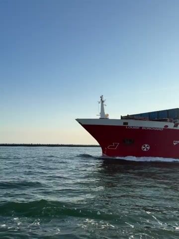 A red and white cargo ship named "LOUISE BORCHARD" is sailing on the water. The ship is loaded with blue shipping containers. The sky is clear and blue. The water is choppy with white foam visible around the ship's hull. The perspective is from the water level, looking towards the ship as it moves from left to right.