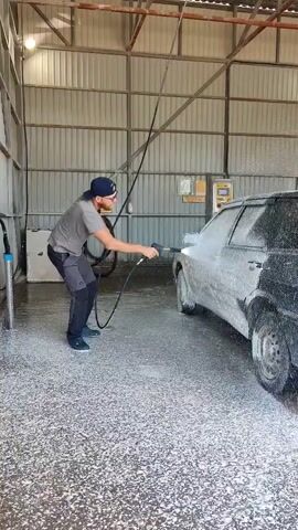 The video shows a man washing a black and silver hatchback car inside what appears to be a commercial car wash. The man is wearing a grey t-shirt, black pants, a dark blue baseball cap and glasses. He's standing on a soapy floor, holding a high-pressure washer nozzle, spraying the car with foam. The car is covered in white foam, and the floor is also coated with it. The car wash facility has corrugated metal walls and a metal roof structure. A hose is hanging from the ceiling. There are some utility boxes or panels on the wall behind the car. The car has a license plate with the code "A 966 XE 82." The man is focusing on washing the car's rear and side panels.