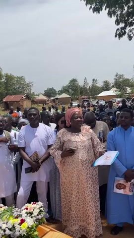 A medium shot shows a crowd of people gathered outdoors under a bright sky, apparently for a funeral. The camera is positioned slightly below eye level, focusing on three people standing near the front. The woman in the center wears a light pink patterned dress and a matching head covering, she holds a smartphone in one hand and a booklet in the other, and is gesturing with both hands as if speaking to the crowd. To her left, a man in a white outfit holds a small booklet and a phone. To her right is a man in a blue robe holding a booklet with a portrait on the cover. A wooden coffin adorned with flowers is in the foreground. The background shows more people and rural buildings with corrugated metal roofs.