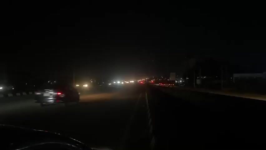 A first-person perspective video taken at night shows a car driving on a highway. Headlights from oncoming traffic illuminate the dark road, creating a hazy glow in the distance. The median of the highway is lined with a black and white barrier, and on the left side of the road, a billboard is visible. The video continues for a few seconds, showing the car moving forward on the highway with cars in front and oncoming traffic.