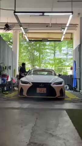 A white Lexus sedan is parked in a garage or workshop. The license plate is visible. A person is standing on the driver's side of the vehicle, apparently cleaning or detailing the car. The space has fluorescent lighting fixtures overhead. Car cleaning supplies are visible along the left side of the shot. Beyond the garage or workshop entrance is some greenery.