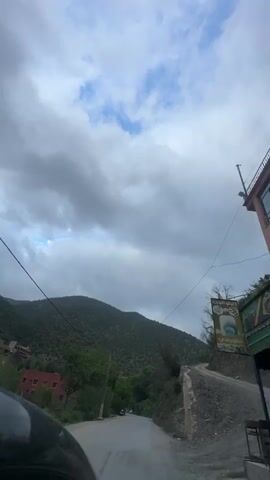 The video shows a first-person perspective from a moving vehicle, likely a car, traveling along a winding road through a mountainous landscape. Two individuals on motorcycles are seen ahead on the road. The scene is set in a rural area with lush green trees and dense vegetation covering the mountain slopes. Power lines crisscross the frame overhead. The sky is overcast with heavy grey clouds, though patches of blue sky are visible in some areas. The road appears to be paved in some sections and has a rough, unpaved shoulder with gravel and rocks on the right side, next to a concrete retaining wall. Some buildings with red roofs are partially visible on the left side, nestled among the trees. The overall impression is of a journey through a scenic, rugged, and verdant mountain pass in Taljarft, Morocco. No visible text is present.