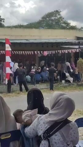 A crowd of people are gathered in front of a building with a covered outdoor area. The building is decorated with red and white banners and pink balloons. Many people are standing and some are sitting on the ground. In the foreground, three people are partially visible from behind, with two of them wearing headscarves. One of these individuals is holding a baby. A motorcycle is parked on the right side of the frame. The sky is overcast.