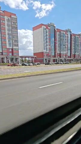 The video is a first-person perspective from inside a vehicle, likely a bus or car, showing a street scene with modern red and white apartment buildings on the right. The road has white lane markings, including red triangle markings for a yield sign. Cars are visible on the road and parked in front of the buildings. The sky is blue with some white clouds. The buildings appear to be multi-story residential structures with many windows, some with balconies. There is a street sign with red and green elements, and another sign with a coat of arms and text that is difficult to read. The camera is slightly tilted and the bottom of the frame shows part of the vehicle's interior, possibly a window frame and blinds. The video appears to be moving forward, showing a continuous view of the street.