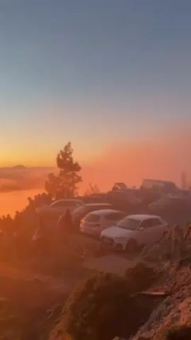 The video shows a sunrise above the clouds from a high vantage point. Several cars are parked in the foreground, near a lone tree. People are gathered on the rocky edge, silhouetted against the orange glow of the rising sun. The sky transitions from a light blue to an orange hue near the horizon, where the sun is emerging. Clouds blanket the lower area, creating a sea of orange mist.