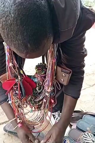 A person, presumably a vendor, is crouched down displaying their merchandise. They are wearing a dark-colored shirt and have numerous necklaces and beaded items hanging around their neck. Additionally, they have what appears to be a wallet or small bag attached to one of the necklace strands. In front of them are an assortment of sandals. The person is tying something, possibly adjusting one of the items for sale. The environment appears to be an outdoor marketplace or street vending area, as the ground is textured and uneven.