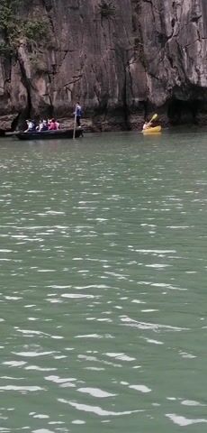 The video shows a body of water near a rocky shoreline with visible trees or vegetation. In the water, there are two boats; one is a long, narrow boat filled with people. A person standing in the boat appears to be guiding it along the edge of the rocky shoreline. The second boat is a yellow kayak with a single person paddling it. The water surface is textured with ripples, and the sky suggests an overcast or slightly hazy day. The overall impression is that of a scenic tour or recreational activity in a natural environment.