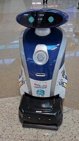 A stationary robot stands in an indoor space, likely a lobby or airport terminal, evidenced by the polished granite-like flooring and blurred people walking in the background. The robot has a shiny blue and silver body, a black base and an oval-shaped head with illuminated green eyes that change. The robot's body features graphics including images of hands holding light bulbs and the words “serveU” in both English and Arabic.