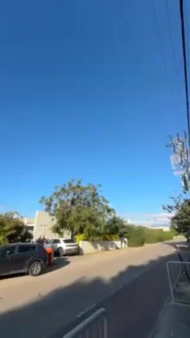 The video shows a suburban street on a bright, sunny day with a clear blue sky. There are several parked cars on the left side of the street, including a dark gray SUV and a white SUV. The street is paved and has sidewalks on either side. There are trees and bushes lining the street, and some houses are visible behind the greenery. Power lines run overhead. The camera pans slowly to the left, capturing more of the street and the surrounding houses.