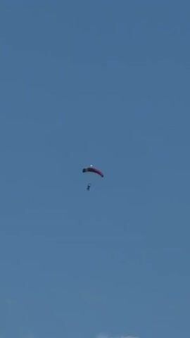 A paraglider is captured mid-flight against a clear blue sky. The paraglider, predominantly red, is in the upper center of the frame, appearing to descend. A small, dark human figure is visible beneath the paraglider, suspended by lines, indicating a successful deployment of the parachute. The surrounding environment is the expansive sky, with a hint of white cloud at the bottom edge of the frame. The camera angle is from below, looking upwards at the paraglider.