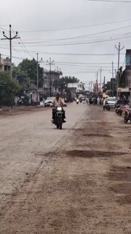 A man rides a motorcycle down a dirt road. He is wearing a light-colored shirt and dark pants. There are other motorcycles and cars on the road. There are buildings and power lines on either side of the road. The sky is overcast.