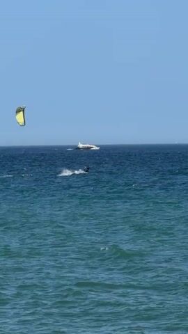 A wide shot shows a clear blue sky over a blue ocean. In the distance, a white boat is visible on the water. Closer to the foreground, a person is kiteboarding, being pulled across the water by a yellow kite. The waves are moderate, and the water is a deep blue. The scene appears to be a sunny day at the beach.