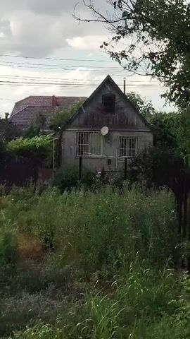 A still shot video showing a small, old house with a steep roof and a satellite dish. The house is surrounded by overgrown vegetation. In the background, there is another house with a red roof and some power lines. The sky is cloudy.