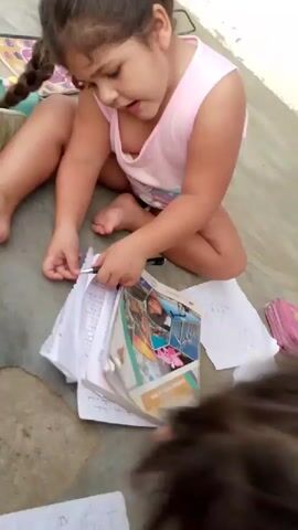 A young child with brown hair in two pigtails wearing a pink tank top sits on a concrete floor surrounded by papers. The child is holding a pen and looking at a book with colorful images. Another young child with brown hair in a bun, wearing a green and pink shirt, is visible in the background, sticking their tongue out. The children appear to be drawing or doing schoolwork.