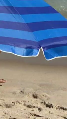The video shows a beach scene. Focus is on the lower bodies of people walking along the shoreline. One individual walks under a blue and white striped beach umbrella, partially obscured. A woman in a black bikini and a person in patterned swim trunks walk alongside. The background includes the beach sand and the ocean.