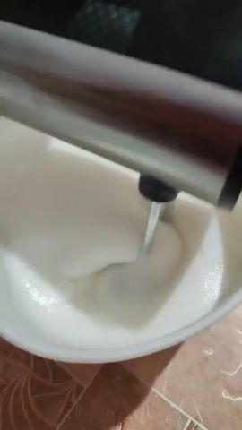 A person is using a black and silver hand mixer to whip a white, frothy liquid in a white bowl. The mixer is partially submerged in the liquid, and the beaters are visible, creating swirls and peaks. The bowl appears to be made of plastic and has a handle. The surface the bowl is resting on is tiled with a pattern of brown and tan squares. In the background, a person's arm is visible, wearing a white shirt. The overall lighting is dim. The audio consists of the whirring sound of the mixer and a voice speaking Russian, stating "We are making a cake."