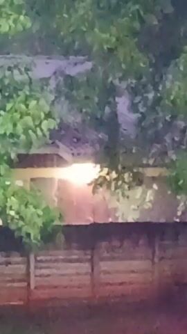 The video shows a view through foliage towards a distant wall. Behind the wall, there is a glowing light source, partially obscured. The light source appears to be illuminating the area behind the wall with an orange tint. The wall itself is constructed of concrete blocks. The scene has a slightly blurry and indistinct quality. The foliage is green. The video appears to be taken during twilight or night.