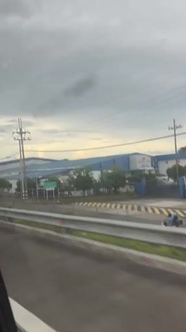 A cloudy sky looms over a row of industrial buildings. A paved road runs in the foreground, bordered by a metal guardrail. Utility poles with numerous wires stretch across the frame. The buildings appear to be warehouses or factories with blue roofs and white walls. Several trees are planted in a row between the road and the buildings. Traffic appears to be moving on the road.