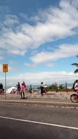 A sunny day by the beach, with a wide view of the sky, sea, and sand. The beach is filled with people, sun umbrellas, and chairs. People are walking, standing, and sitting, some in groups. A yellow caution sign, streetlights, and a traffic light are visible along the road. The road is empty, with a white and red crosswalk. The sea has small islands in the distance. The sky is bright blue with white clouds.