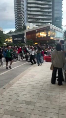 A man in a green and white soccer jersey and black shorts, holding a microphone and a beer, dances on a paved walkway in front of a crowd. He is wearing a blue backpack. Behind him, a Sephora store sign is visible, along with a tall building and a restaurant with illuminated windows. Other people in the crowd are also wearing green and white, some are sitting, and some are standing and cheering. A few individuals are dancing. The ground is paved with light-colored square tiles.