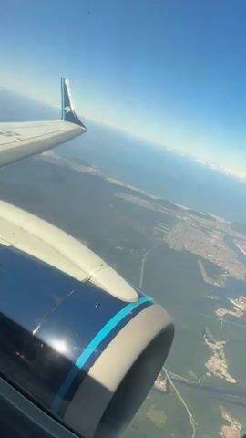 A shot from an airplane window shows the airplane wing and engine in the foreground. Below is a vast landscape with a city, water, and greenery. The city appears to be coastal, with buildings lining a body of water that extends towards the ocean. The sky is clear and blue, and the horizon shows a faint line of clouds or distant land.