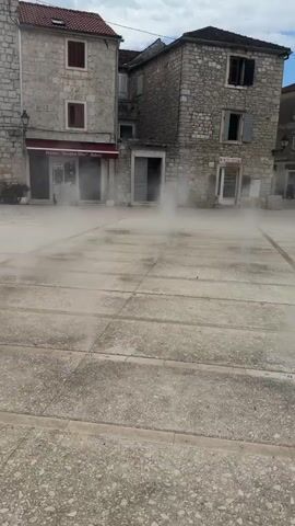 The video shows a public square with stone buildings and pavement. Mist or fog is present, obscuring the lower portions of the buildings and the ground in the center of the frame. A person walks from right to left across the square in the background. The sky is overcast.