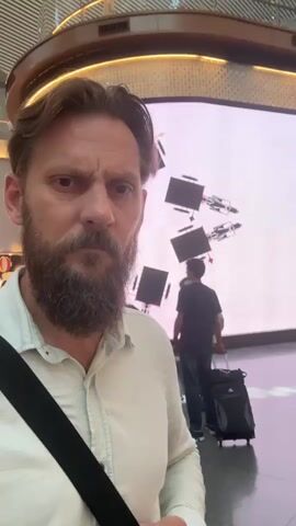 A man with a beard takes a selfie in what appears to be an airport. He is wearing a light-colored shirt and a black strap across his chest. Behind him, a large digital screen displays a pattern of repeating geometric shapes and what appear to be Louis Vuitton logos. Other people are visible in the background, including one person pulling a suitcase and another standing near a store entrance with a "LOUIS VUITTON" sign. The lighting is bright, reflecting off the polished floor. The man's expression is one of mild surprise or confusion.