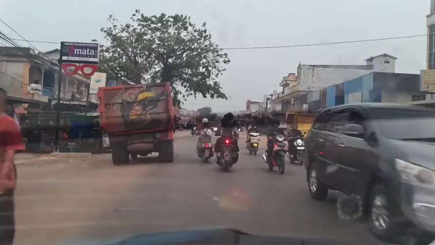 A street scene in a town or city. The camera is looking forward from inside a vehicle, possibly a car, as it moves along a two-lane road. The road is paved and appears to be in moderate condition. On the left side of the road, there is a large red dump truck with graffiti on its side. Further down the left side are several buildings, some appearing to be shops or small businesses, with awnings and signs. A large tree with dense foliage is prominent on the left side, casting some shade on the road. On the right side of the road, there are more buildings, some multi-story, with various advertisements and structures. A yellow truck is parked on the right.

The road is populated with many motorcycles and scooters, with a few cars and other vehicles interspersed. Several people are riding on the motorcycles, some with passengers. The general atmosphere suggests a moderately busy urban or semi-urban environment, possibly during the day or early evening given the lighting. The sky appears overcast or hazy. There are no obvious signs of distress or emergency. The overall impression is that of ordinary daily traffic and commercial activity.