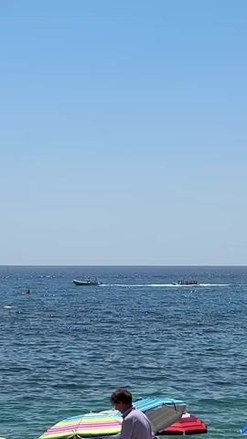 A person wearing a purple shirt stands on a beach, partially obscured by beach umbrellas. In the distance, the ocean is visible under a clear blue sky. A boat tows several people on a long inflatable raft across the water, leaving a white wake behind it. Another smaller boat is visible further to the left. A few other people are in the water or on flotation devices.