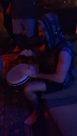 A man with a beard and a hooded vest sits on a couch, intensely playing a handheld drum. The lighting in the room shifts colors, occasionally casting a pink or blue hue. Around him, other people stand and walk, some barefoot, while a patterned rug sits on the floor beneath the couch. The man is the central focus, rhythmically striking the drum with both hands.
