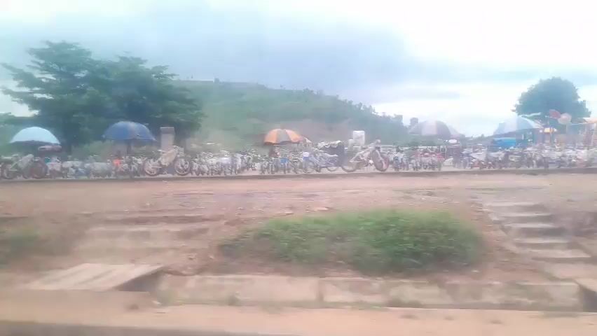The video captures a roadside scene under a cloudy sky, with a line of motorcycles parked along a dirt road. Several umbrellas are set up, providing shade over the motorcycles. In the background, a lush green hill rises, dotted with trees. A few cars, including a white van and a black sedan, are visible. One person is seen walking in the foreground. A small wooden bridge crosses a ditch in front of the parked vehicles. The overall impression is that of a busy market or gathering place.