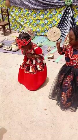 Two young girls in traditional outfits dance outside. The first girl wears a red dress with a patterned bodice and a matching beaded crown. The second girl wears a black skirt with tulle, a patterned bodice with lion faces, and a beaded necklace. A man in a white outfit and blue hat is behind them. The dancing takes place in front of a colorful backdrop and wooden bowls with round objects. A small wooden chair sits to the side. A sign on the building above reads "God's Favourite Academy" and lists "Daycare" and "Nursery."