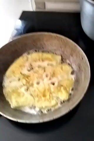 The video shows two cooking scenes. The first scene, timestamped 00:00, is a close-up shot of eggplant slices being fried in a pan. The eggplant is coated in a batter and sizzles in hot oil, with bubbles forming around the pieces. The second scene, from timestamp 00:01 to 00:05, shows a plate piled high with golden-brown fried eggplant pieces. In the background of this scene, a large pot filled with a bubbling, light-brown liquid containing pieces of food, possibly meat and vegetables, is visible.