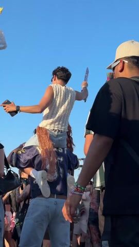 The video captures a vibrant crowd at an outdoor event under a clear blue sky, likely a music festival in Indio, United States of America, during the daytime.

Several people are visible in the crowd. In the foreground, from a slightly elevated perspective, a person wearing a black t-shirt, a black backpack, and a light-colored cap stands with their back to the camera. To their right, another person in a green and white striped shirt and a black cap is also visible from behind.

The camera then pans to focus on a person in the foreground wearing a stylish white lace long-sleeved top over a red bra, with long, dark, curly hair. They accessorize with a gold chain belt featuring red circular ornaments and a black bracelet on their left wrist. A black and white bandana is tied to their waist. This person is seen swaying and briefly raising their hand.

In the background, throughout the video, a person in a white knit top can be seen sitting on someone's shoulders, raising one arm in the air, possibly holding a phone, and holding a patterned fan with the other hand. The person carrying them wears a blue and white sports jersey. Other blurred figures of people are also visible in the background, contributing to the lively atmosphere.

A lanyard with "Heineken" and "EMERGENCY" visible on it can be seen at the beginning of the video, worn by someone in the foreground. People appear to be enjoying themselves, cheering, and dancing to music that can be heard in the background.