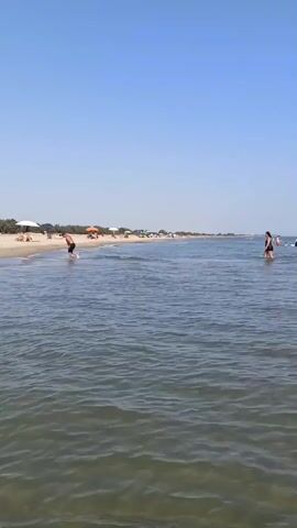 The video shows a wide, calm body of water with a sandy beach in the background. The sky is clear and blue. Several people are visible on the beach, some are standing, and some are sitting under umbrellas. A few people are in the water. One man is walking in the water towards the left of the frame. A woman is standing in the water on the right side of the frame. The overall scene depicts a peaceful day at the beach.