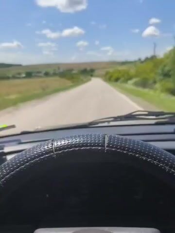 The video shows the view from inside a car, looking forward. The steering wheel is visible in the foreground, with a textured grip. The road ahead is paved and curves slightly to the left. The landscape on either side is rural, with fields and trees under a blue sky with scattered white clouds. The driver's side window is partially open, showing a side mirror and the passing scenery. The video appears to be a first-person perspective of someone driving through the countryside on a sunny day. There is a sound of music playing, with lyrics that are partially audible.