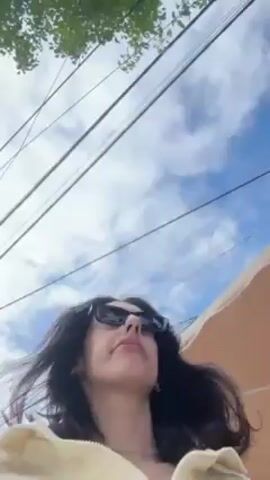The video shows a woman outdoors, likely in a tropical area. She is wearing sunglasses and a light-colored jacket with a small Polo logo on the front. She is seen running her hand through her hair. The background includes trees, buildings, and a road. Decorations, possibly red ribbons, are visible in the trees, adding a festive touch. The sky is partly cloudy.