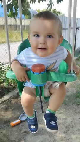 A young boy is sitting in a green swing. He is wearing a white and blue striped t-shirt and blue shoes. He is smiling and appears to be enjoying himself. The swing is outdoors, and there are trees and a fence in the background.