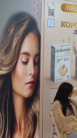 The video captures scenes from an indoor exhibition or trade show, likely in Vynnyky, Ukraine, given the visible Ukrainian text. The event appears to be focused on beauty, cosmetics, and health products.

Several people, predominantly women, are seen engaging with various display booths. In the initial shots, a large promotional banner features a woman's face alongside text in Ukrainian that reads "ЛІНІЙКА α-БІСЕЛЕН: ВАШ КОРОННИЙ ВИБІР ДЛЯ ВОЛОССЯ!" (LINE α-BISELEN: YOUR CROWN CHOICE FOR HAIR!). Product boxes labeled "α-Біселен" and "α-Біселен DHT" are prominently displayed, suggesting hair supplements or care products. One woman is seated near this display, and another walks past.

Another segment shows a well-decorated display stand adorned with fresh flower bouquets, including white roses, purple delphiniums, and pink peonies. Small product boxes are arranged on shelves, set against a backdrop banner featuring a woman wearing a crown, reinforcing the "crown choice" theme.

Further into the video, the scene shifts to other exhibition booths. Multiple women are seen interacting with products on sleek white display stands that house numerous cosmetic bottles and jars. Banners with English text such as "GROUP SOLUTIONS," "HD COSMETIC EFFICIENCY," and "PROFESSIONAL SKIN SOLUTIONS" are visible, indicating different brands or product lines. One counter clearly displays the text "ELEGANT GROUP." Attendees are observed bending over displays, examining products, and browsing the offerings. The overall atmosphere is professional and engaging, with bright indoor lighting.