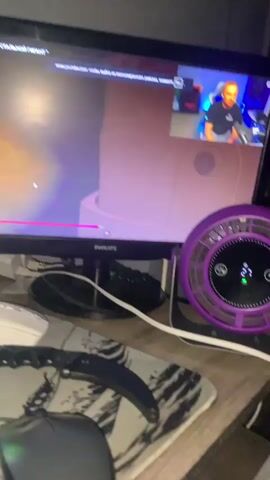 The video shows a computer monitor displaying a YouTube video. The YouTube video shows an animated scene with a character with large blue eyes and an orange shirt. The YouTube video is captioned in Russian. In the top right corner of the monitor, there is a smaller window displaying a live video feed of a person sitting in front of a computer. The desk in front of the monitor has a keyboard, a mouse, a knife with a karambit-style handle, and a purple fan.